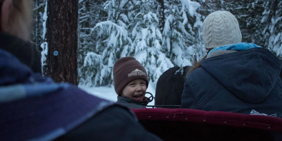 little boy with a hat on smiling while riding on a sleigh through the snow covered forest