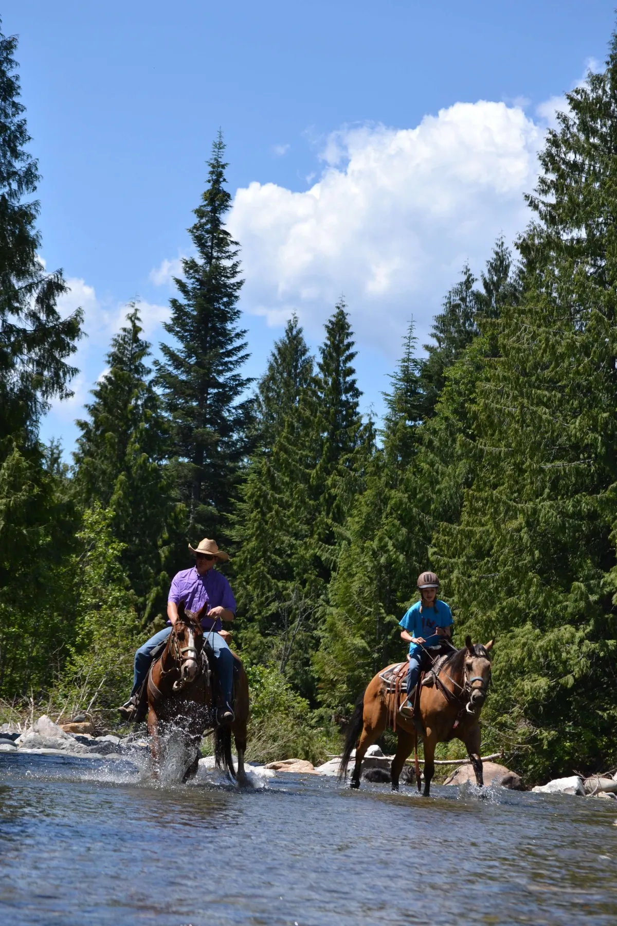Two people on an adventure horseback riding across a river in the forest.