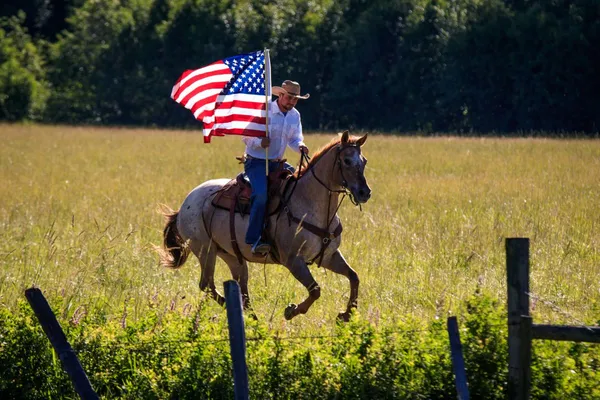 Cowboy running an Appaloosa Gelding through a green field while holding a waving American flag