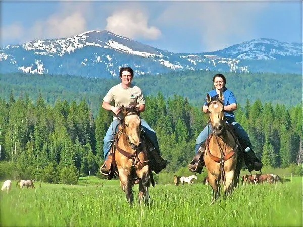 Isaac and Libby riding buckskin horses through a meadow with snowy mountains behind