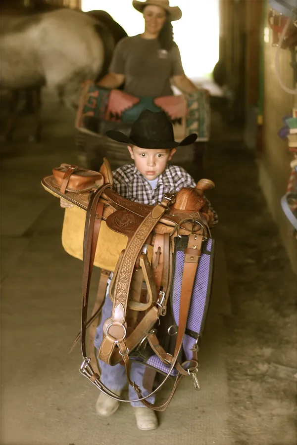 A small boy in a cowboy hat carrying a saddle as big as he is through the barn