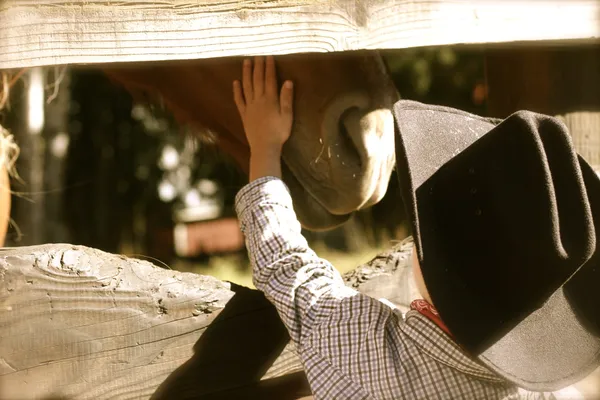 A child in a cowboy hat reaches up to pat a horse's nose