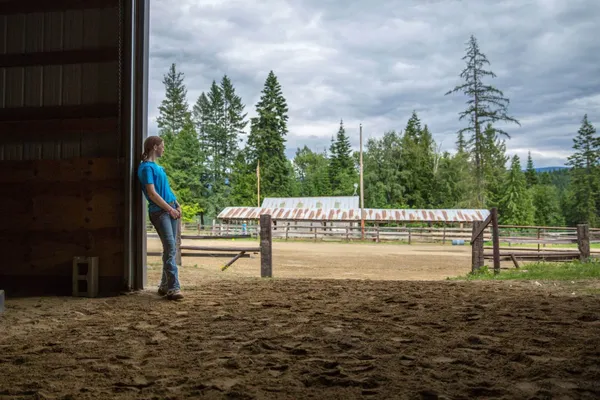 Girl leaning against a barn looking out at an arena and a rusting barn roof
