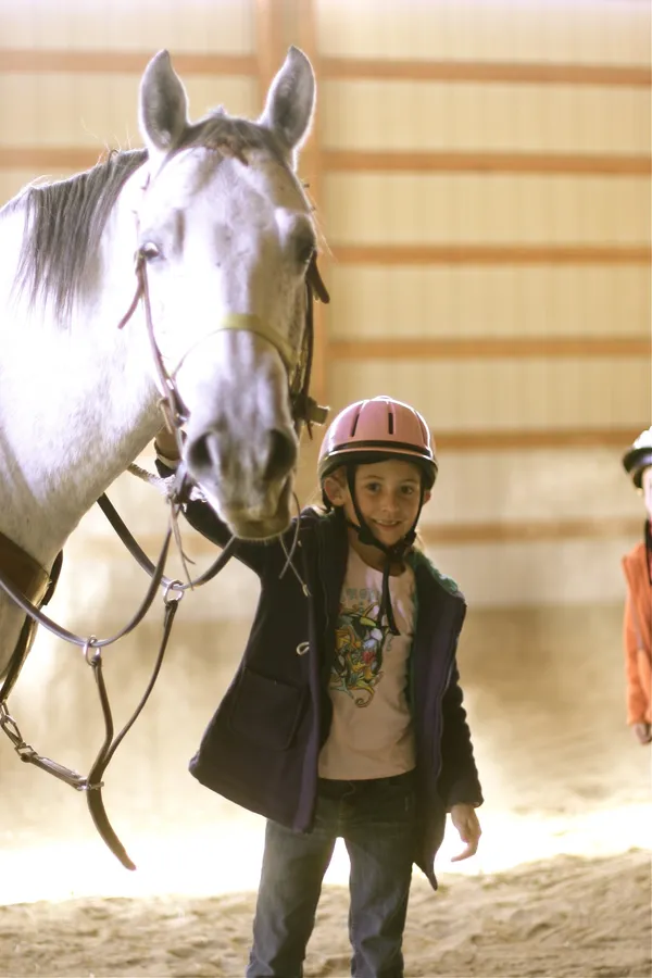 A young rider in a helmet stands beside a grey horse in the indoor arena