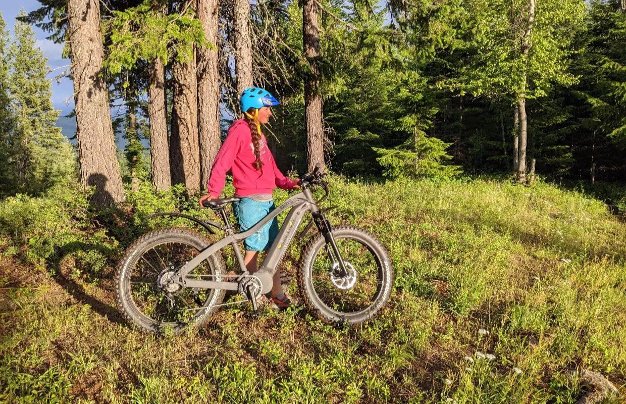 A person wearing a blue helmet and pink hoodie stands with a fat-tire electric mountain bike in a sun-drenched Idaho forest clearing.