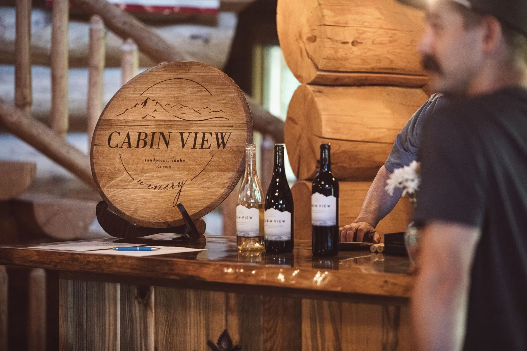 Bottles of Cabin View Winery wine on the lodge bar beside a round wooden sign reading "Cabin View Winery, Sandpoint, Idaho"