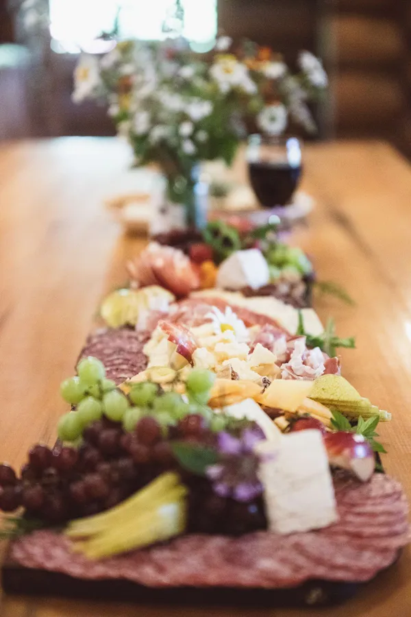A long charcuterie board down a wooden table, with a jar of wildflowers and a glass of red wine at the far end