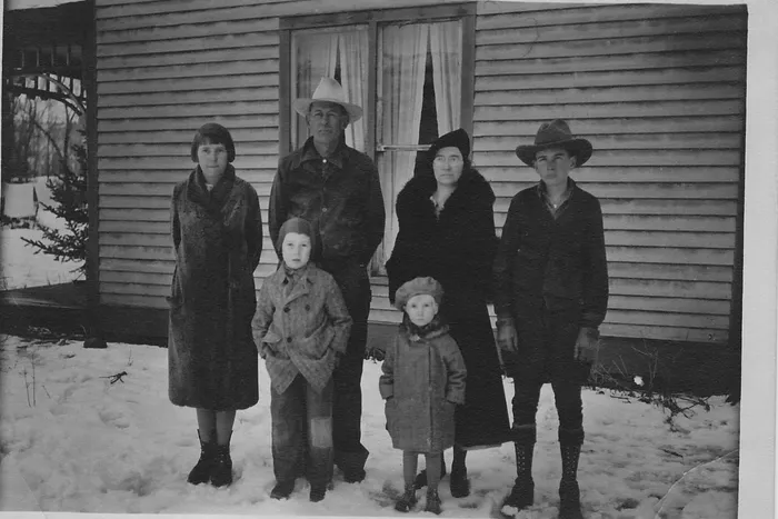 Old black and white photo of a husband, wife and four kids in coats and hats outside a house in the snow