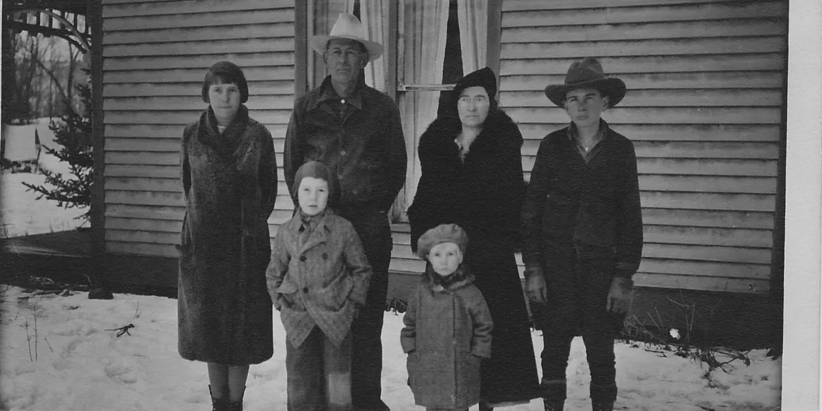 western history Old black and white historical photo of husband, wife and 4 kids standing with coats and hats on outside of a house in the snow