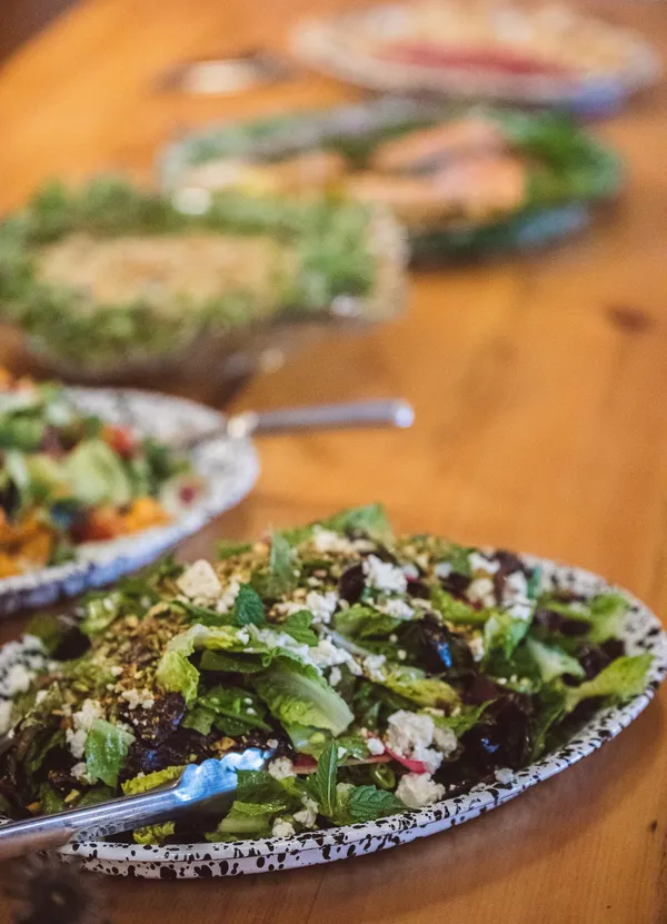 A green salad with feta and pistachios in a speckled enamel dish, with more salads down the table