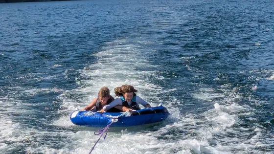 Two laughing kids in life jackets riding a blue tube towed behind a boat on a lake, with forested hills behind