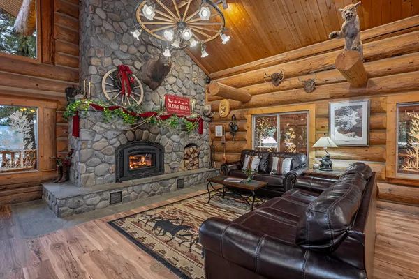 Great room river rock fireplace with an elk mount and a mantel dressed for Christmas, leather couches on a horse-pattern rug, and a cougar mount on a log beam