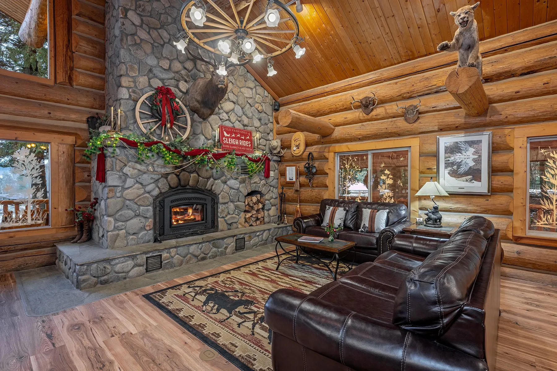 Great room river rock fireplace with an elk mount and a mantel dressed for Christmas, leather couches on a horse-pattern rug, and a cougar mount on a log beam