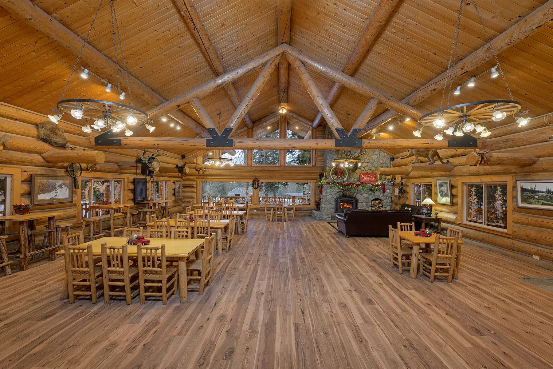 Great room with log trusses, wagon-wheel chandeliers, rows of log dining tables and chairs, and a river rock fireplace at the far end
