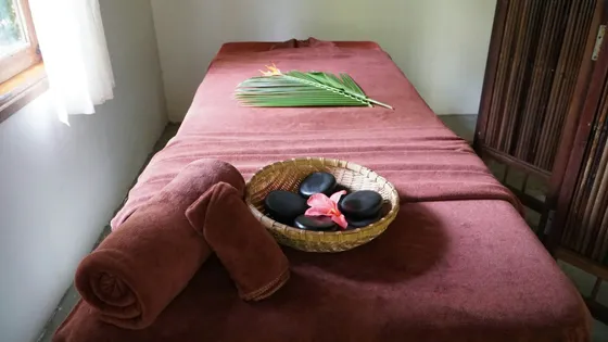 A massage table with rolled towels and a basket of smooth dark stones beside a sunlit window