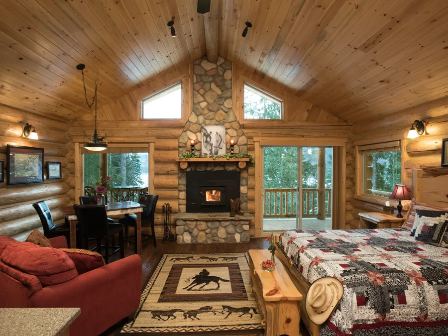 Inside the Meadow View Cabin: a river rock fireplace with a fire lit, a quilted queen bed, a small dining table and a door out to the deck
