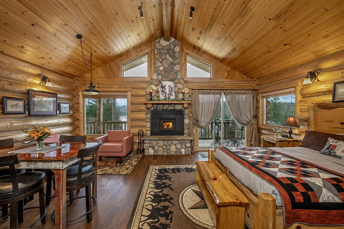 Main room of the Meadow View Cabin, with a river-rock fireplace, a log bed with a patchwork quilt, a dining table and doors to the deck