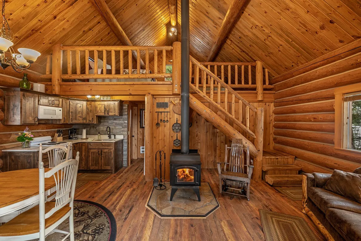 Main room of the Settler Cabin, with a wood stove burning, the kitchen, and log stairs up to the open loft