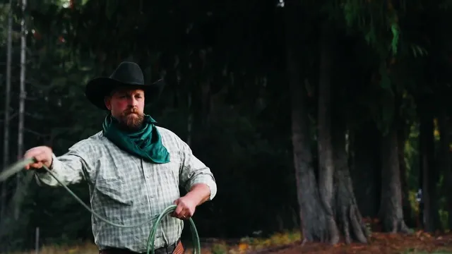 A cowboy in a black hat and green scarf swings a rope at the edge of the pines