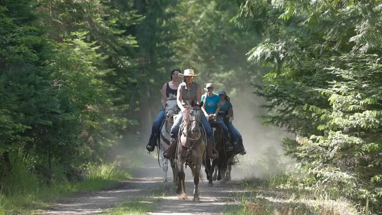 Riders come down a dusty forest trail toward the camera, the lead rider in a straw cowboy hat