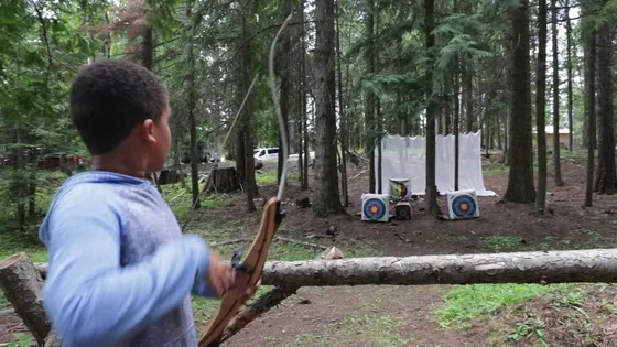 A boy holding a traditional bow looks down the archery range toward targets in the trees
