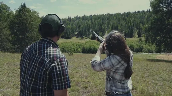 A woman aims a shotgun over a field while an instructor wearing ear protection stands beside her