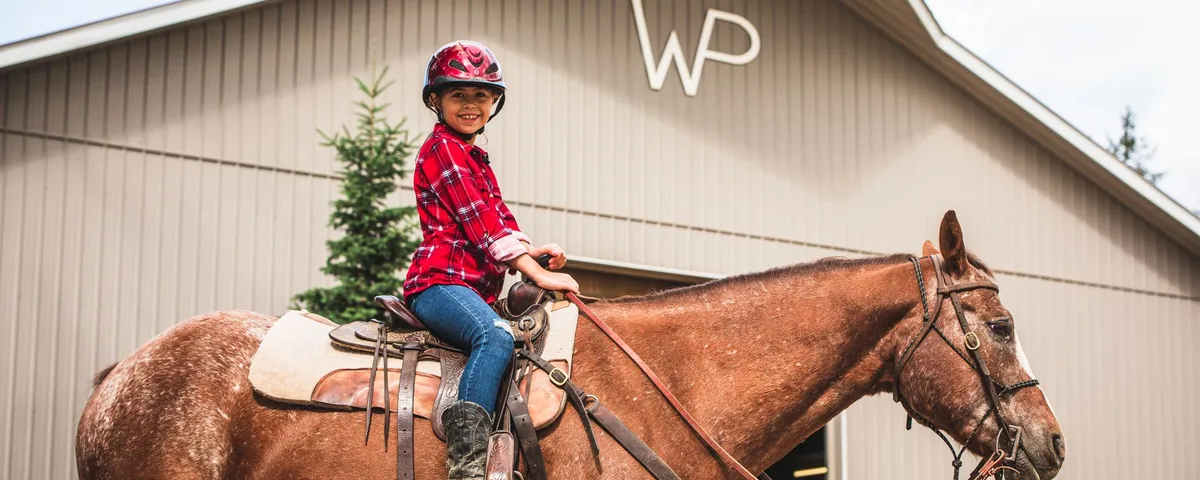 Smiling young rider in a red plaid shirt and helmet on a chestnut horse in front of the barn with the WP brand