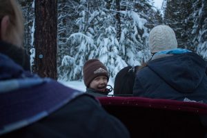 little boy with a hat on smiling while riding on a sleigh through the snow covered forest