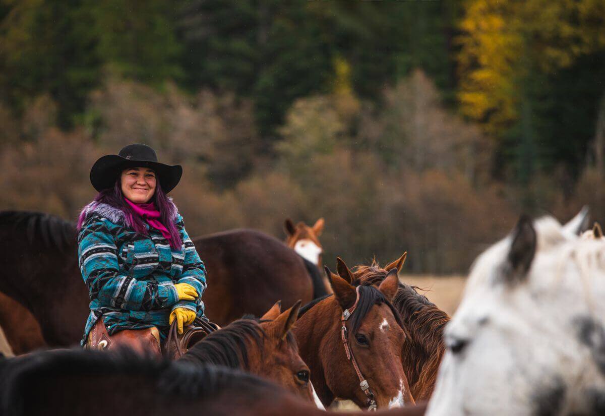 Cowgirl smiling and sitting on ranch horse in a field of horses and fall colors