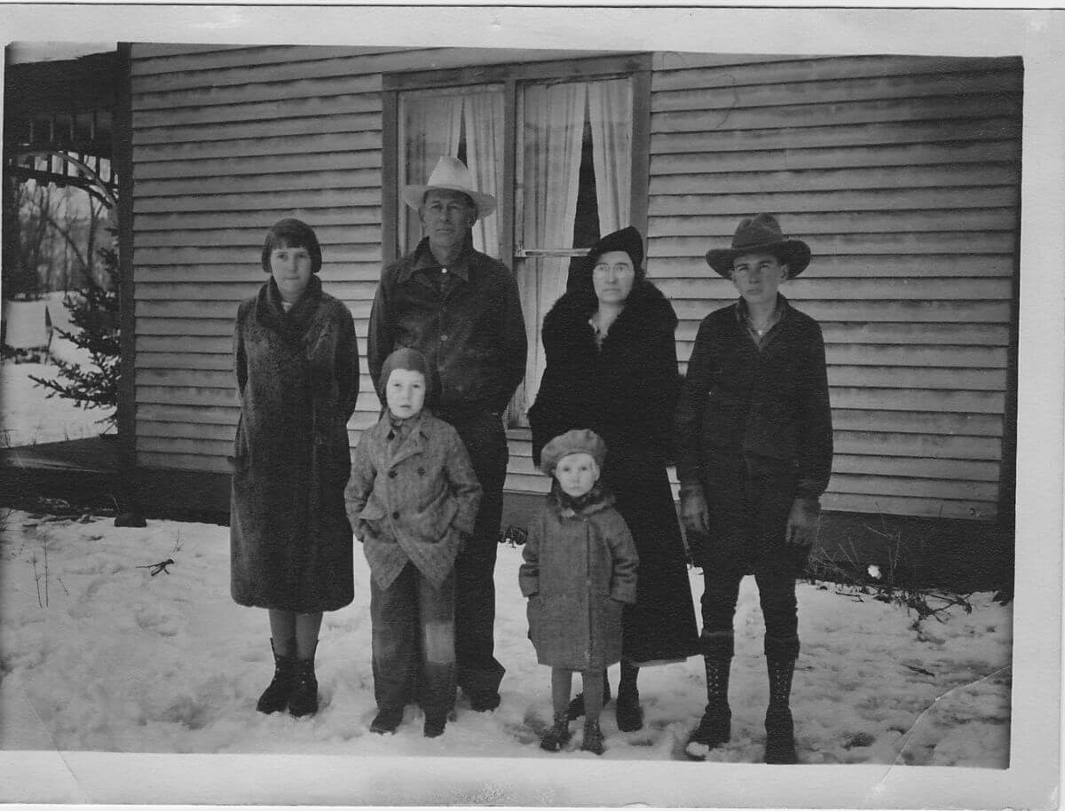 old black and white photo of a family of 6 standing in front of an old house in the snow