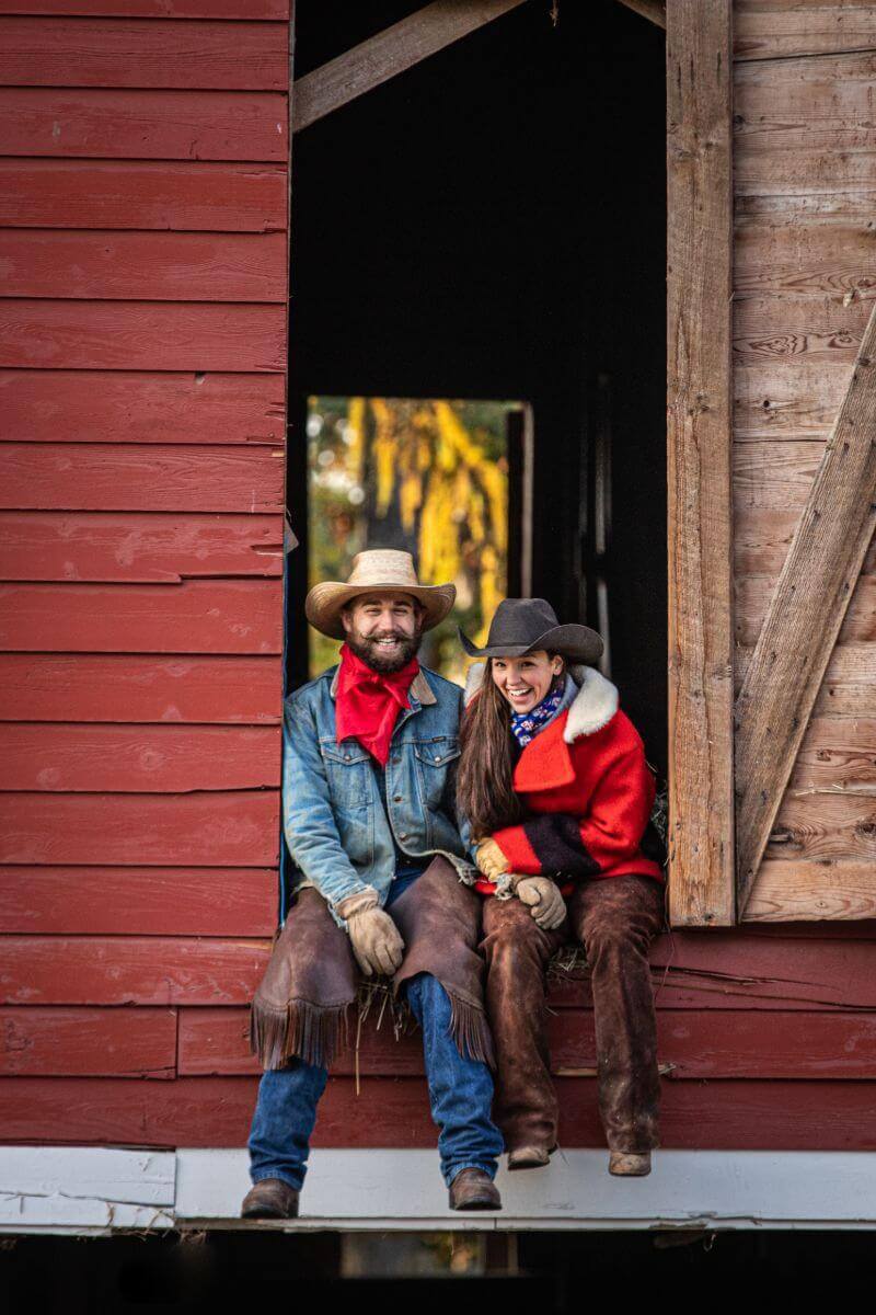 couple wearing cowboy hats sits in the doorway of a red barn